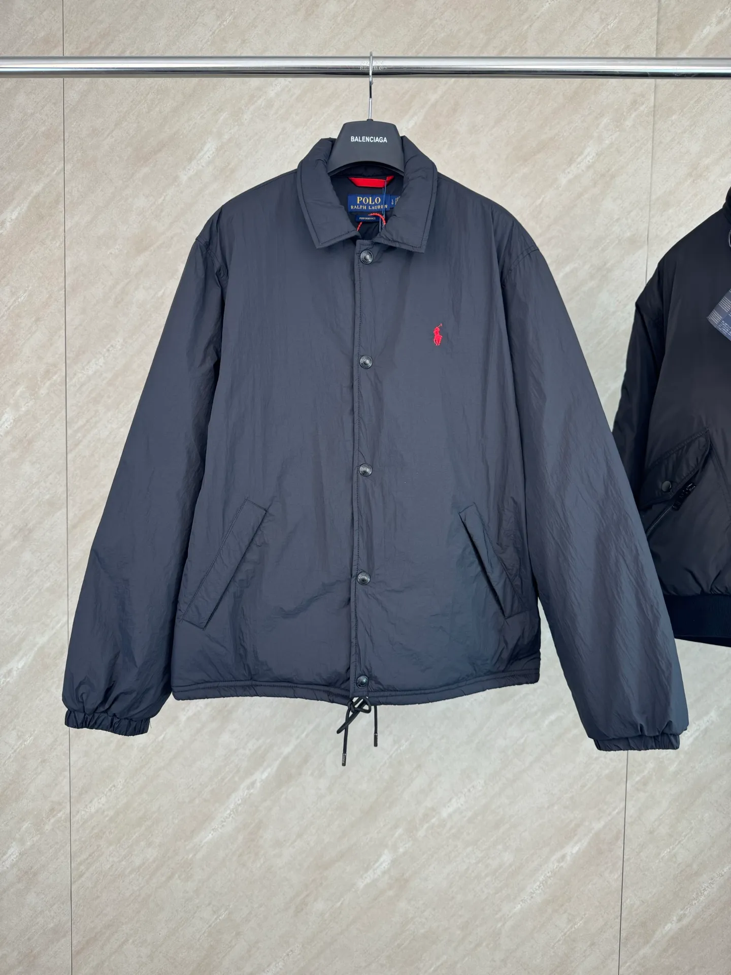 P670 25FW Ralph Lauren red label light cotton jacket (coach jacket) size s m l xl Fabric procurement: a combination of pragmatism and classic texture Material: high-density nylon taffeta Features: density is about 210T-300T, smooth and slightly matte surface, excellent windproof and waterproof performance (with DWR water-repellent treatment), strong wrinkle resistance and easy care, in line with the positioning of American casual "durable and practical".  Color: Classic dark black, using vat dyeing process (dye penetrates into the fiber), the color fastness reaches ISO level 4 or above to avoid fading or color transfer after washing.  2. Lining fabric Material: Lightweight cotton-polyester blended lining (65% Polyester + 35% Cotton) Function: Reduce friction between the fabric and inner clothing, improve wearing smoothness; blended material has both breathability and durability to avoid the stuffy feeling of pure nylon lining.  Version and cutting process Version: Classic Coach Jacket, loose version, slightly dropped shoulder line (shoulder width is 1-2cm wider than the standard version), length to the hips, and slightly tapered straight sleeves, taking into account both freedom of movement and casual style.  Embroidery technology: Polo horse logo on the left chest: flat satin stitch embroidery (satin embroidery), embroidery thread: 100% polyester high color fastness embroidery thread (the color number is consistent with the brand’s standard red color card), stitch density 12-15 stitches/cm, the outline of the horse logo is clear, and the details of the rider and horse are burr-free.  Embroidery positioning: precise positioning through the template (12cm from the neckline and 8cm from the placket) to ensure left and right symmetry and centering.  Topstitching technology: The seams of the body are made of double-needle flat seams (needle pitch 3cm/10 needles), the stitches are smooth and straight, the stitching color is dark black (the same color as the fabric), and the stitch width is 0.5cm, which enhances firmness while forming simple decorative lines.  1. Main body splicing, splicing of the front placket and body: The placket is made of double-layer folding + flat seam splicing, with a thickness of about 0.3cm to ensure that the buttons are not easily deformed when fixed; the edges of the placket are cut with a hot knife to avoid burrs.  Sleeve panels are spliced ​​to the body: the armholes are spliced ​​with round-head keyhole machine seams + flat seams. After splicing, the armholes have a natural curvature and no sense of tightness during movement; the height of the sleeves is about 8-10cm, which is in line with the loose characteristics of casual jackets.  Main button: black urea resin button, matte surface treatment, buttonholes are hand-made keyholes (6 strands of cotton thread, keyhole density 8 stitches/mm), the edges of the eyelets are smooth and without burrs.  Collar buttons: The first button on the collar is a hidden button (diameter 12mm), which prevents the collar from opening and improves the neatness of wearing.